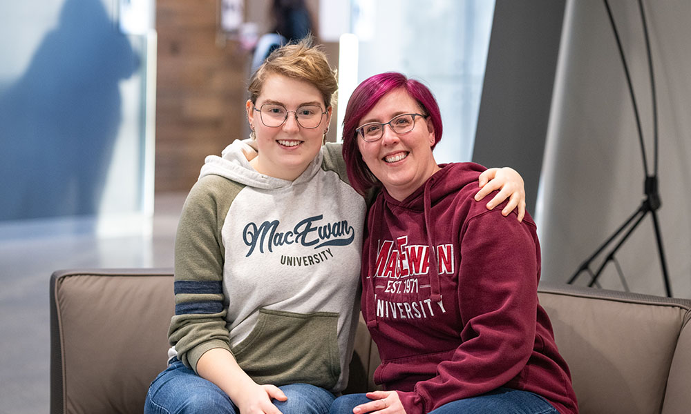 Diana and Heather Eyestone wear MacEwan hoodies and sit on a bench in the SAMU Building